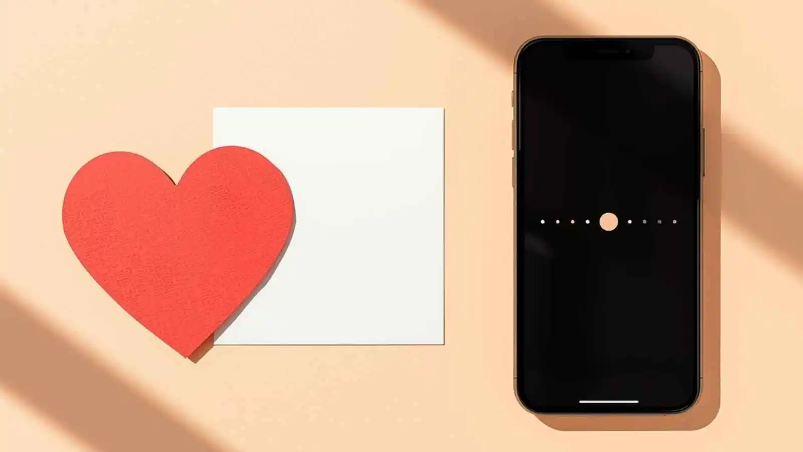A red paper heart beside a blank card and a phone on a bright desk.